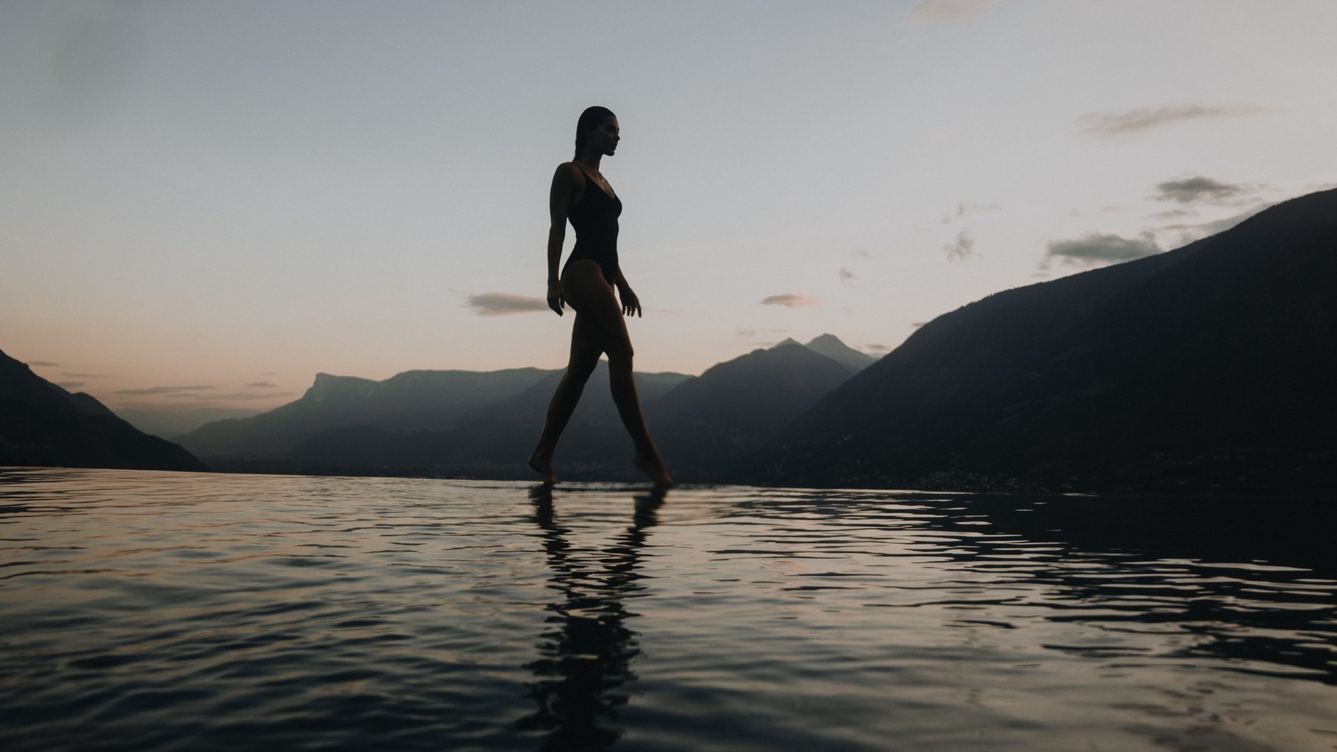 Das wohl schönste Hotel in Dorf Tirol Frau in Badebekleidung geht am Rand eines Pools bei Sonnenuntergang mit Bergen im Hintergrund.